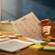 Leraarshanden met beoordelingsformulier en cijferboek op houten bureau, omringd door gekleurde sticky notes in warm middaglicht.